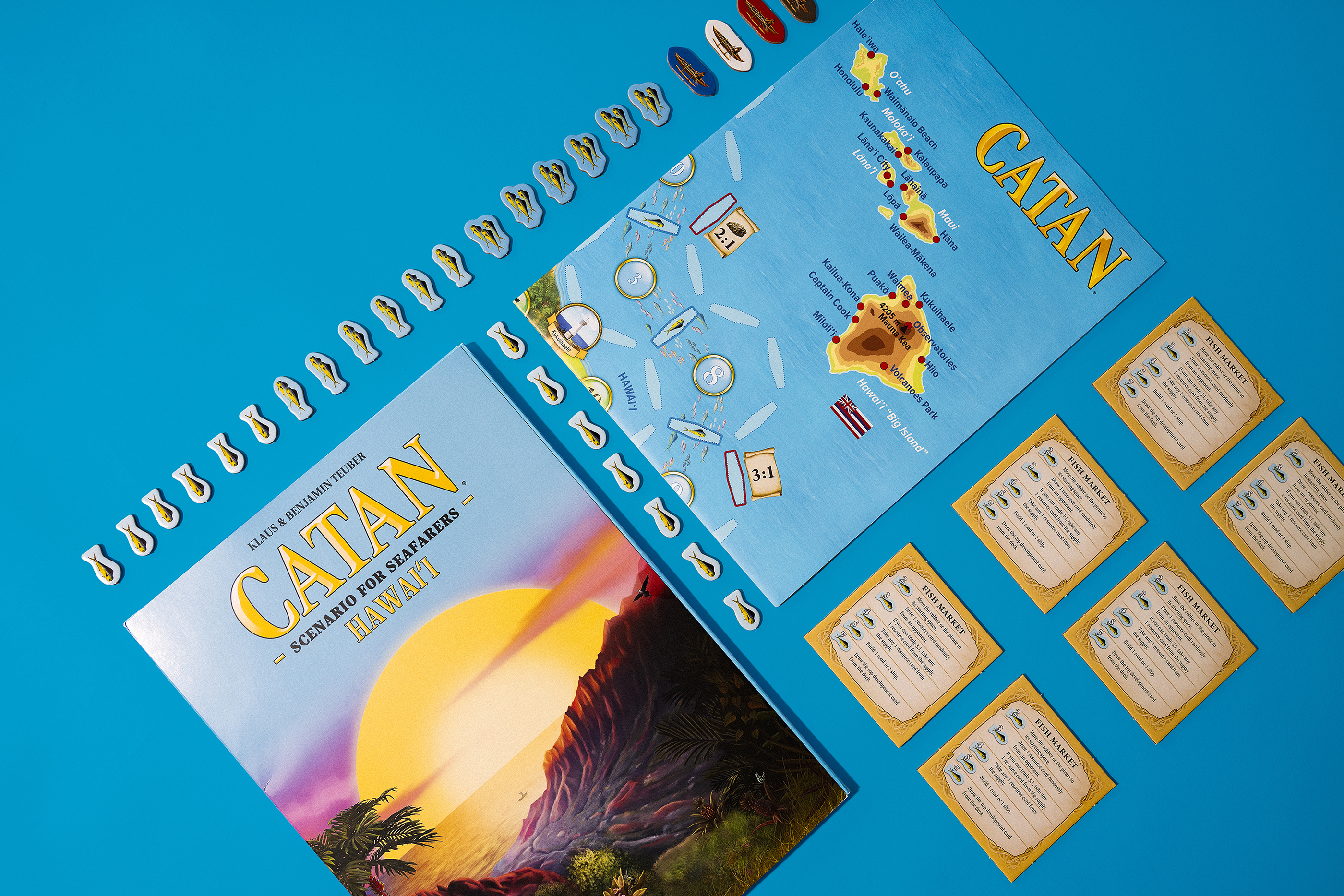 CATAN Hawai'i board, package and pieces laid out on a blue backgroun